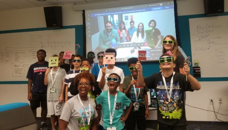 A group of students holding Minecraft toys stands in front of a screen displaying a Skype call with teachers