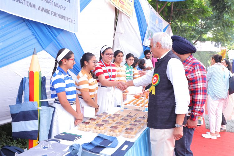 Two dignitaries shake hands with a group of girls standing behind a sales kiosk in a an open-air market.