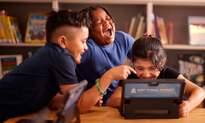 Three students are laughing while using a tablet computer in a classroom