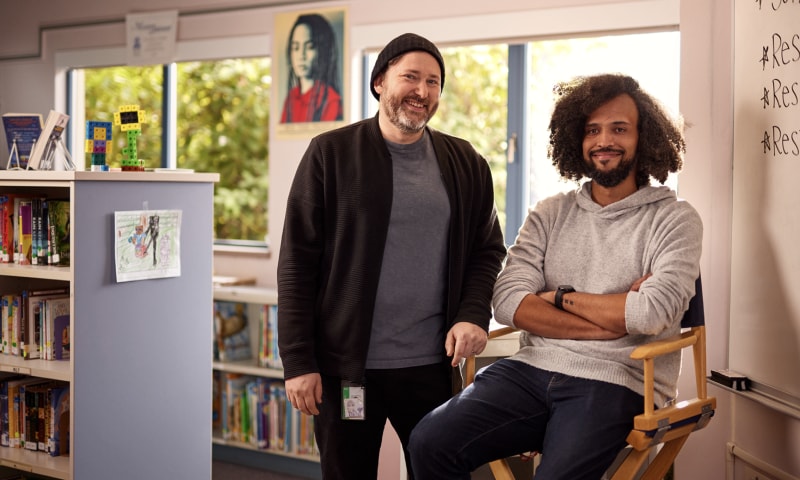 Two people in a school library. A child's drawing of a Minecraft scene hangs from the shelves.