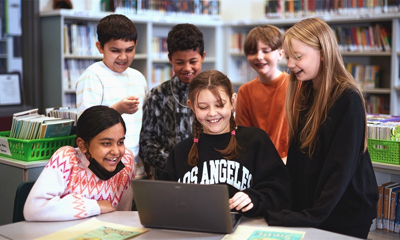 Six children are gathered around a laptop in a school library, smiling and laughing